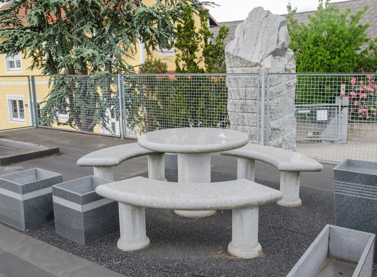Runder Tisch mit drei gebogenen Bänken und Pflanztrögen aus hellem und grauem Stein, dahinter ein großer Steinblock
